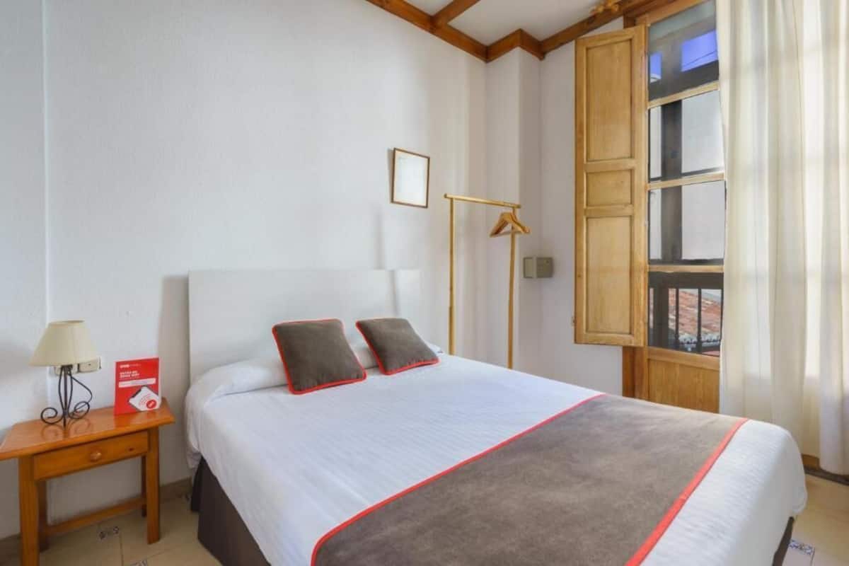 A neatly made bed with a white and grey blanket, a bedside table with a lamp, a framed picture on the wall, and a window with curtains.
