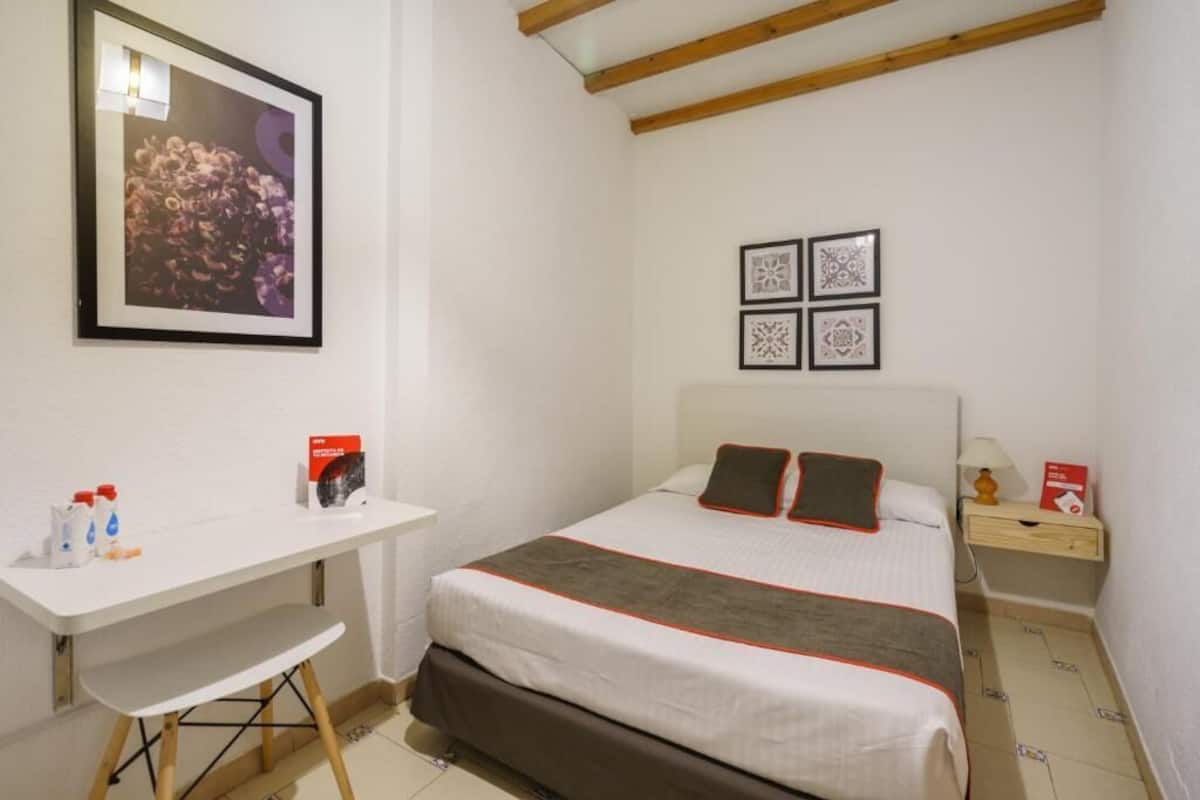 A bedroom with a bed, a desk, and a chair. There are framed pictures on the wall and a small table with a lamp.