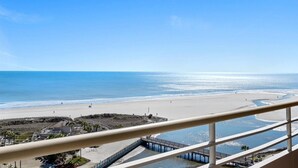 Blick auf einen Strand vom Balkon aus, mit blauem Himmel, Sandstrand und einem sich ins Wasser erstreckenden Steg.