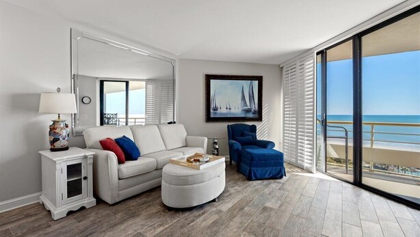 Ein modernes Wohnzimmer mit einem weißen Sofa, einem blauen Sessel und einem kleinen Beistelltisch mit einer Lampe. Ein großes Fenster bietet Blick auf den Ozean.