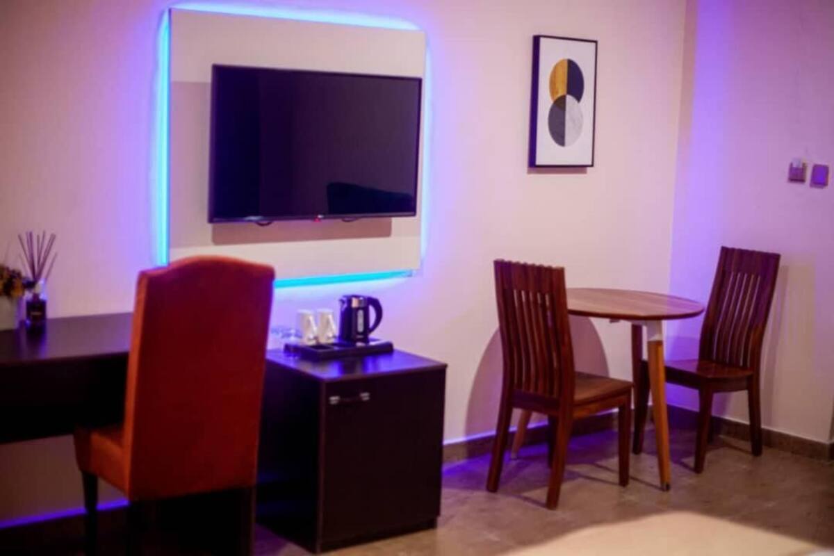 Quarto moderno com televisão de tela plana na parede, uma mesa de centro pequena e duas cadeiras de madeira.