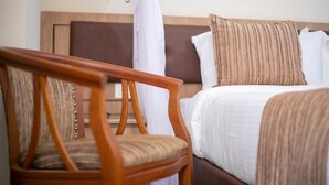 Una habitación de hotel con una silla de madera, una cama con almohadas blancas y a rayas, y una cortina.