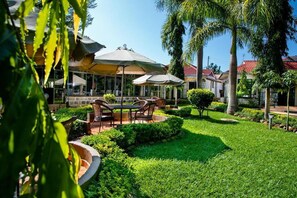 An outdoor seating area with tables and chairs under umbrellas, surrounded by well-maintained greenery and palm trees. - Siaya County Club (Bondo)