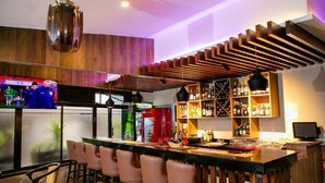 Zona de bar con una barra larga, varios taburetes, una pantalla de televisión y una variedad de botellas y vasos en estanterías.