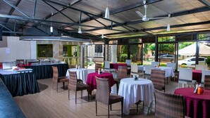 Un comedor interior con mesas redondas y sillas de mimbre, con un techo alto con vigas metálicas y luces colgantes.