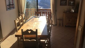 A dining area with a large wooden table and chairs, a view of snow-capped mountains through a large window, and a wooden cabinet with framed pictures on the wall.
