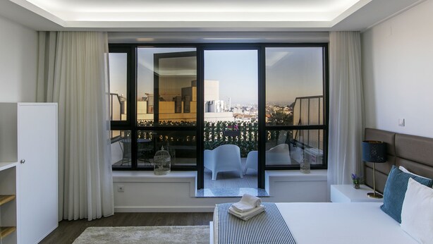 Una habitación de hotel moderna con un ventanal grande, una cama, una mesita de noche y vistas a la ciudad.
