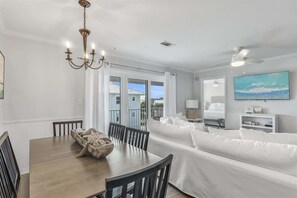 Uma sala de jantar com mesa e cadeiras de madeira, um lustre e um sofá branco com vista para o exterior.