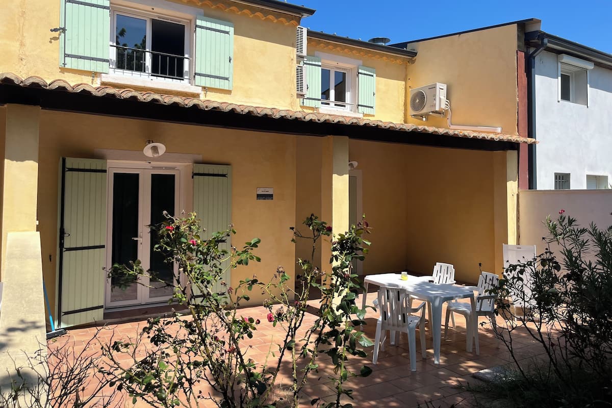 A two-story building with a tiled patio, a dining area with white chairs, and a small garden with plants and flowers.
