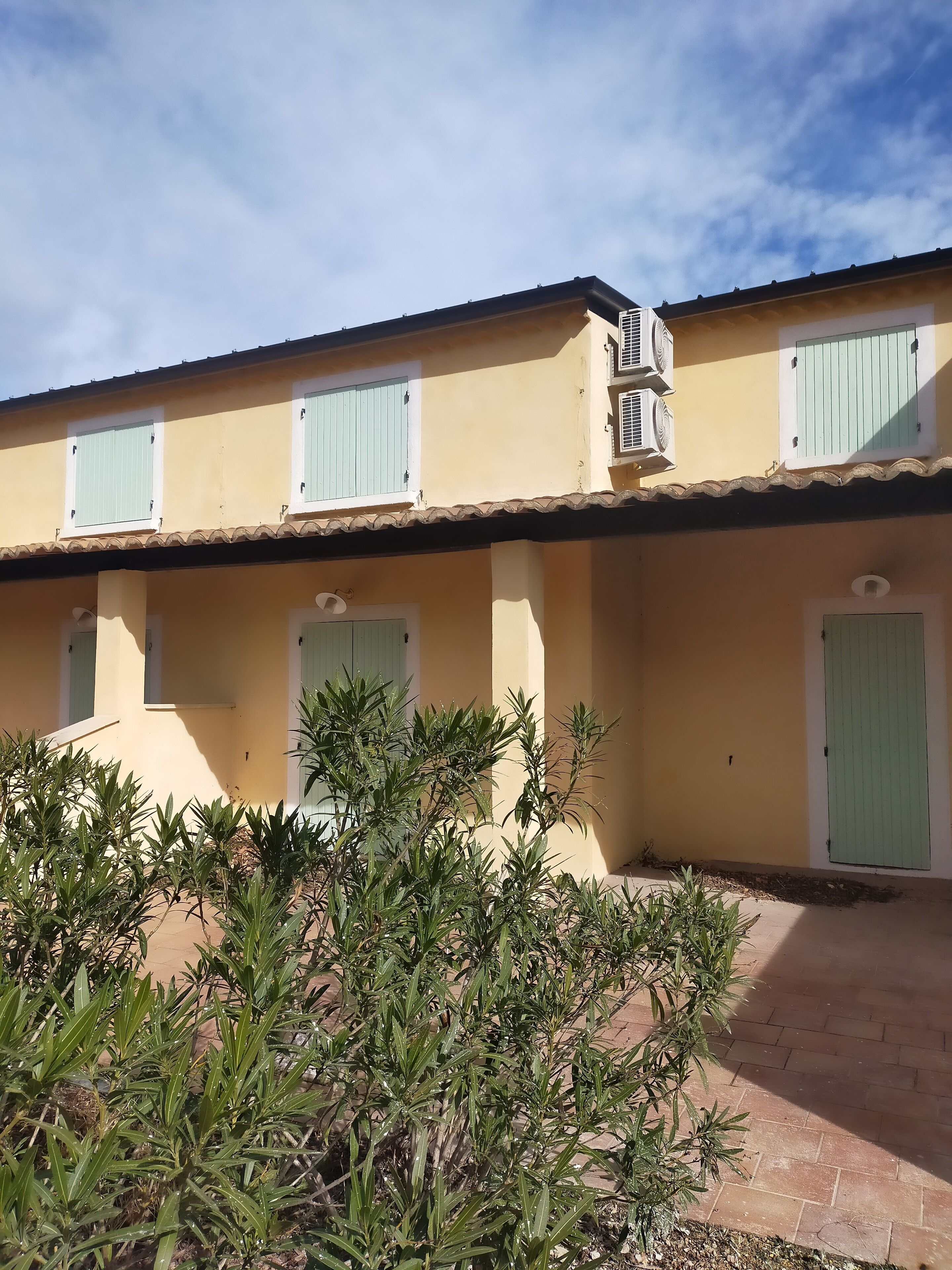 A building with a flat roof, multiple windows with shutters, and a paved entrance area with plants.