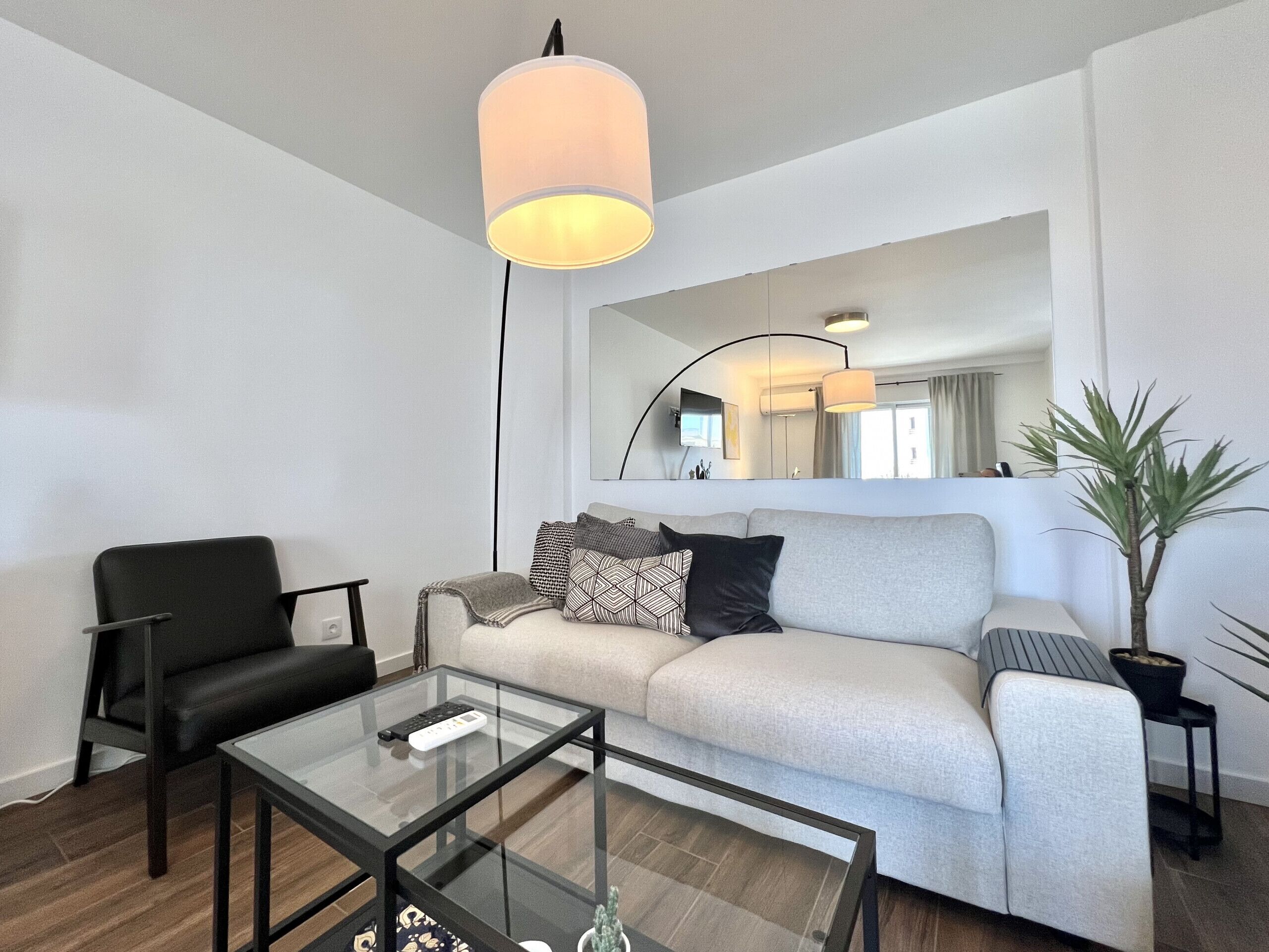 A modern living room with a glass coffee table, a black leather chair, a grey sofa with patterned cushions, a floor lamp, and a potted plant.