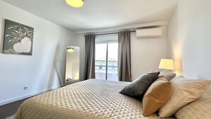 Ein Hotelzimmer mit einem großen Bett, einer Nachttischlampe, einem Fenster mit Vorhängen, einer Klimaanlage und einem Gemälde an der Wand.