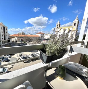 Ein Balkon mit einer Topfpflanze, einem Tisch mit Kissen und Blick auf eine Stadtansicht, darunter ein Kirchturm.