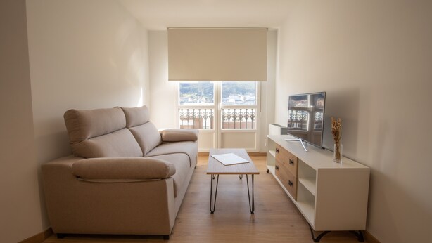 A living room with a sofa, a TV on a stand, a table, and a window with a view of buildings. - Duerming Ría de Viveiro Apartamentos (Viveiro)