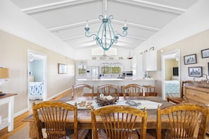 Une salle à manger avec une table en bois, des chaises, un lustre et une vue sur la cuisine.