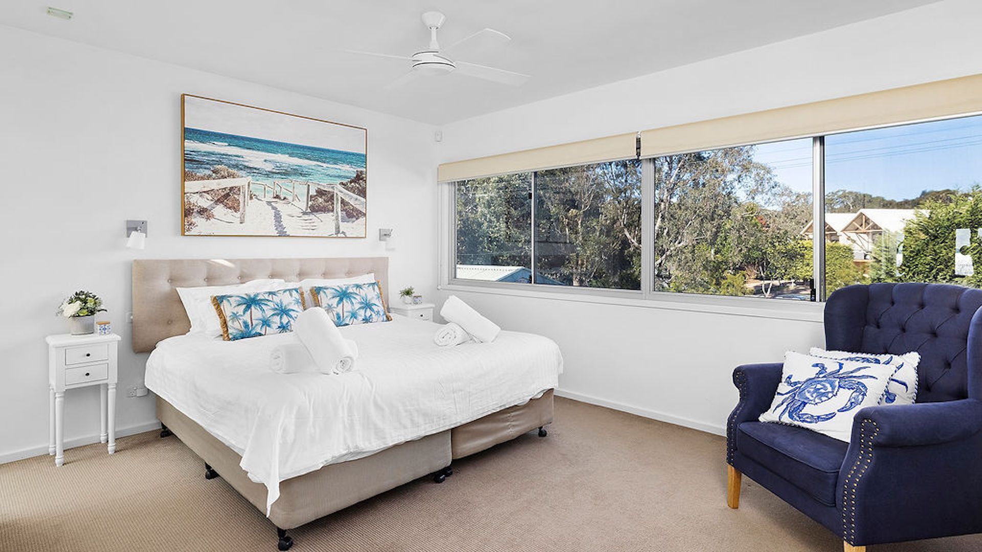 Une chambre à coucher comprenant un lit, une table de chevet, une chaise, un ventilateur de plafond, une fenêtre donnant sur un beau paysage et un tableau accroché au mur.