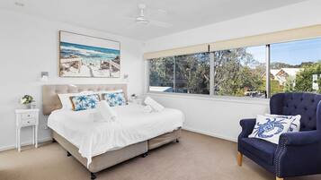 Une chambre à coucher comprenant un lit, une table de chevet, une chaise, un ventilateur de plafond, une fenêtre donnant sur un beau paysage et un tableau accroché au mur.