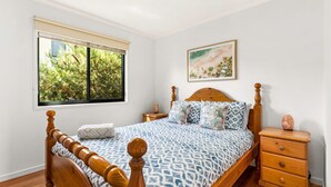 Un dormitorio con una cama de madera, un ventanal con vistas a árboles, un cuadro en la pared y una mesita de noche de madera con una lámpara de sal.