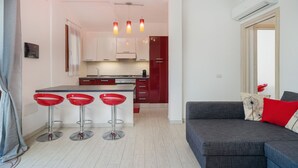 A modern kitchen with white cabinets, a red refrigerator, and a grey sofa with red cushions. There are three red bar stools and a ceiling light fixture.