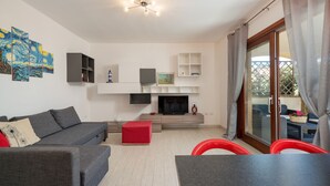 A modern living room with a grey sofa, a red ottoman, a flat-screen TV, and a dining area with a table and chairs.