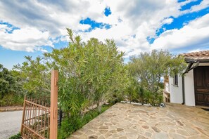 A stone pathway leading to a building with a traditional tiled roof, surrounded by lush greenery and a metal gate.