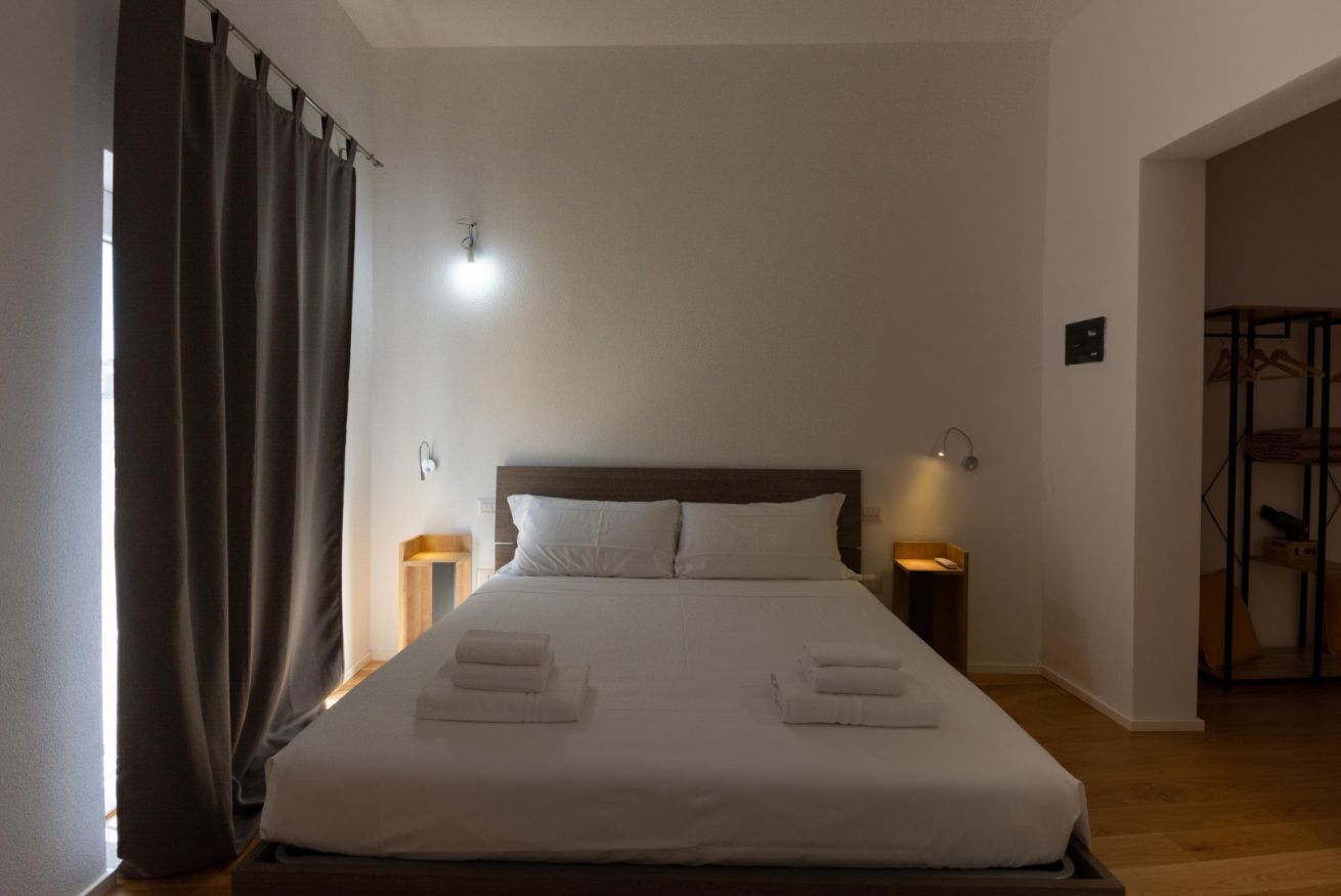 Een net opgemaakt bed met witte lakens en handdoeken, een houten hoofdbord, twee bedlampen en een raam met gordijnen.