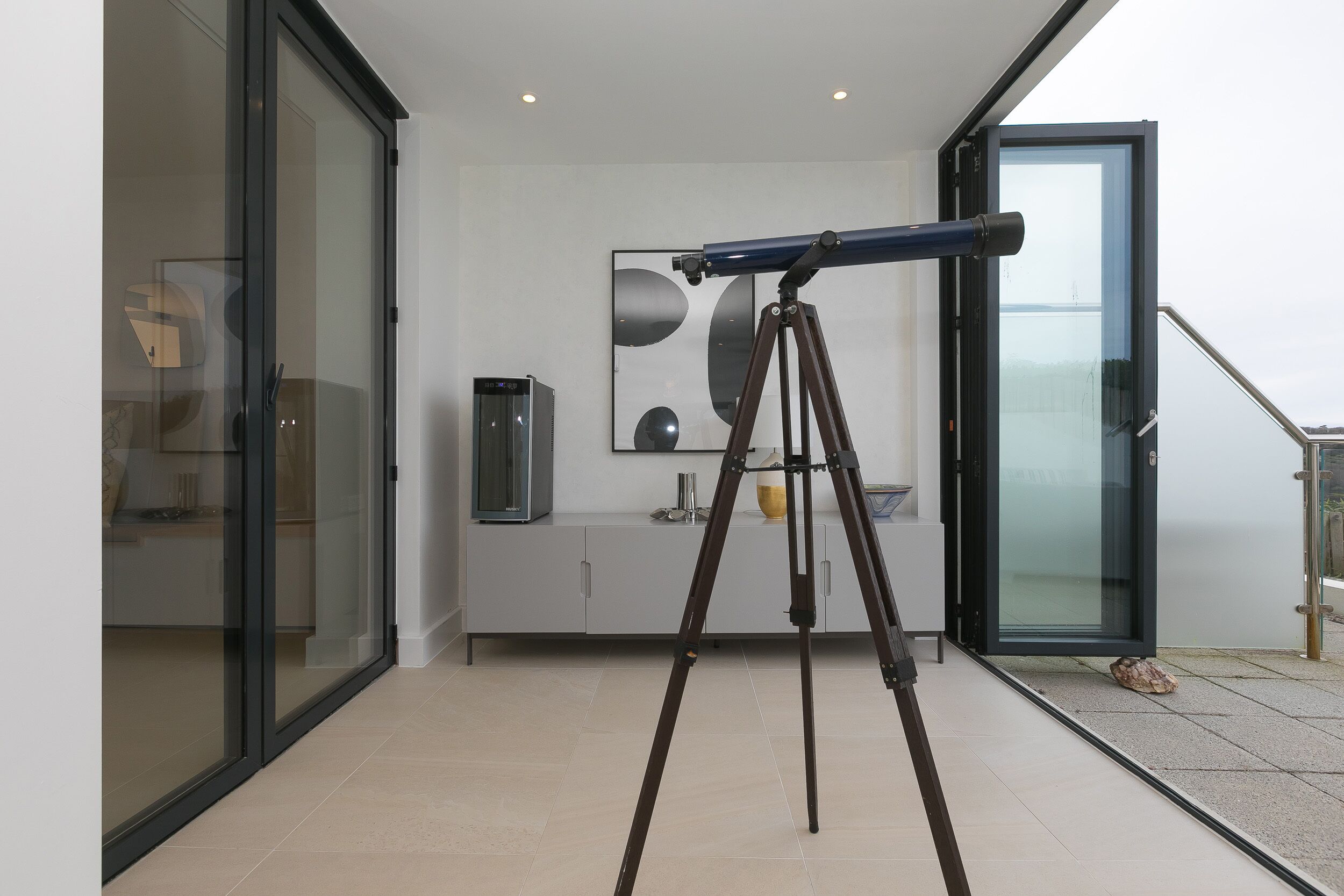 A modern kitchen with a glass door leading to a balcony, a telescope on a tripod, and a decorative wall piece.