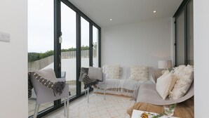 A modern living room with a sofa, armchair, and coffee table, large windows, and a view of the outdoors. - Monowai Heights (St Ives)