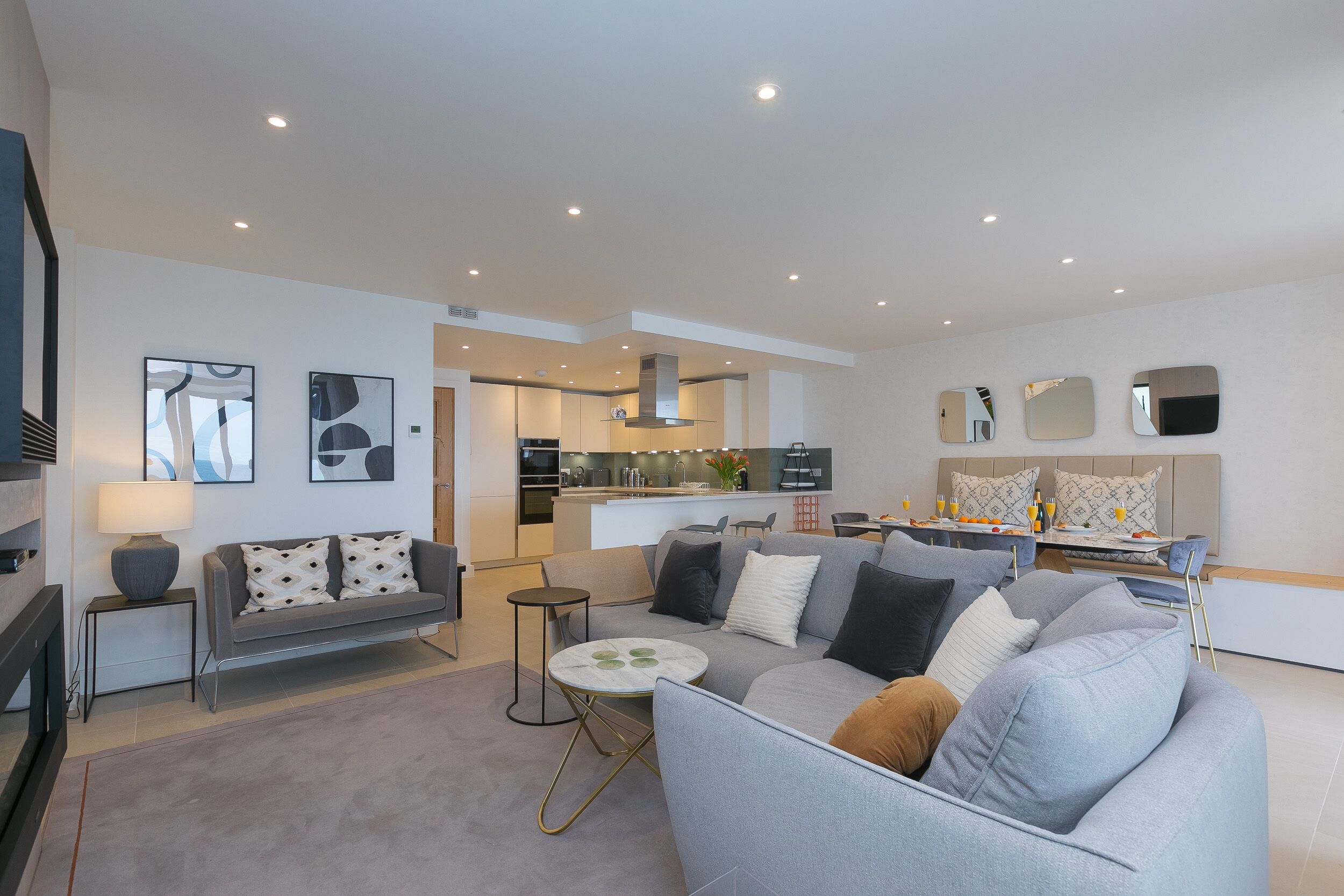 A modern living room with a grey sofa, a coffee table, and a TV. There is a kitchen in the background with a dining area.