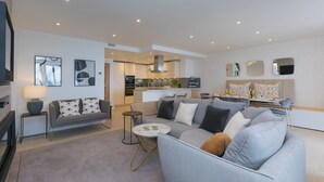 A modern living room with a grey sofa, a coffee table, and a TV. There is a kitchen in the background with a dining area. - Monowai Heights (St Ives)