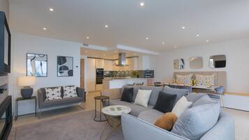 A modern living room with a grey sofa, a coffee table, and a TV. There is a kitchen in the background with a dining area.