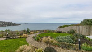 A landscaped garden with a curved pathway, a wooden fence, and a view of the sea. - Monowai Heights (St Ives)