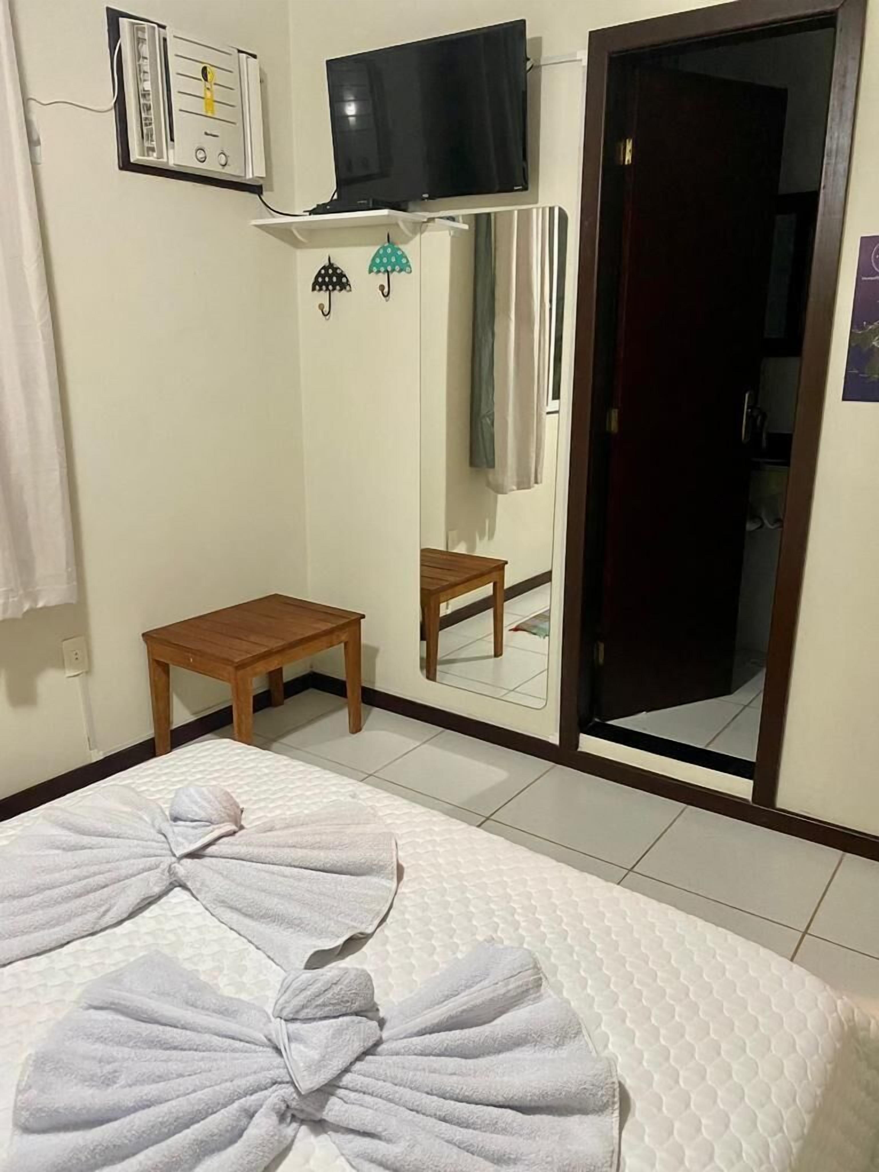 A hotel room with a bed, a small wooden table, a mirror, a TV mounted on the wall, and a bathroom door.