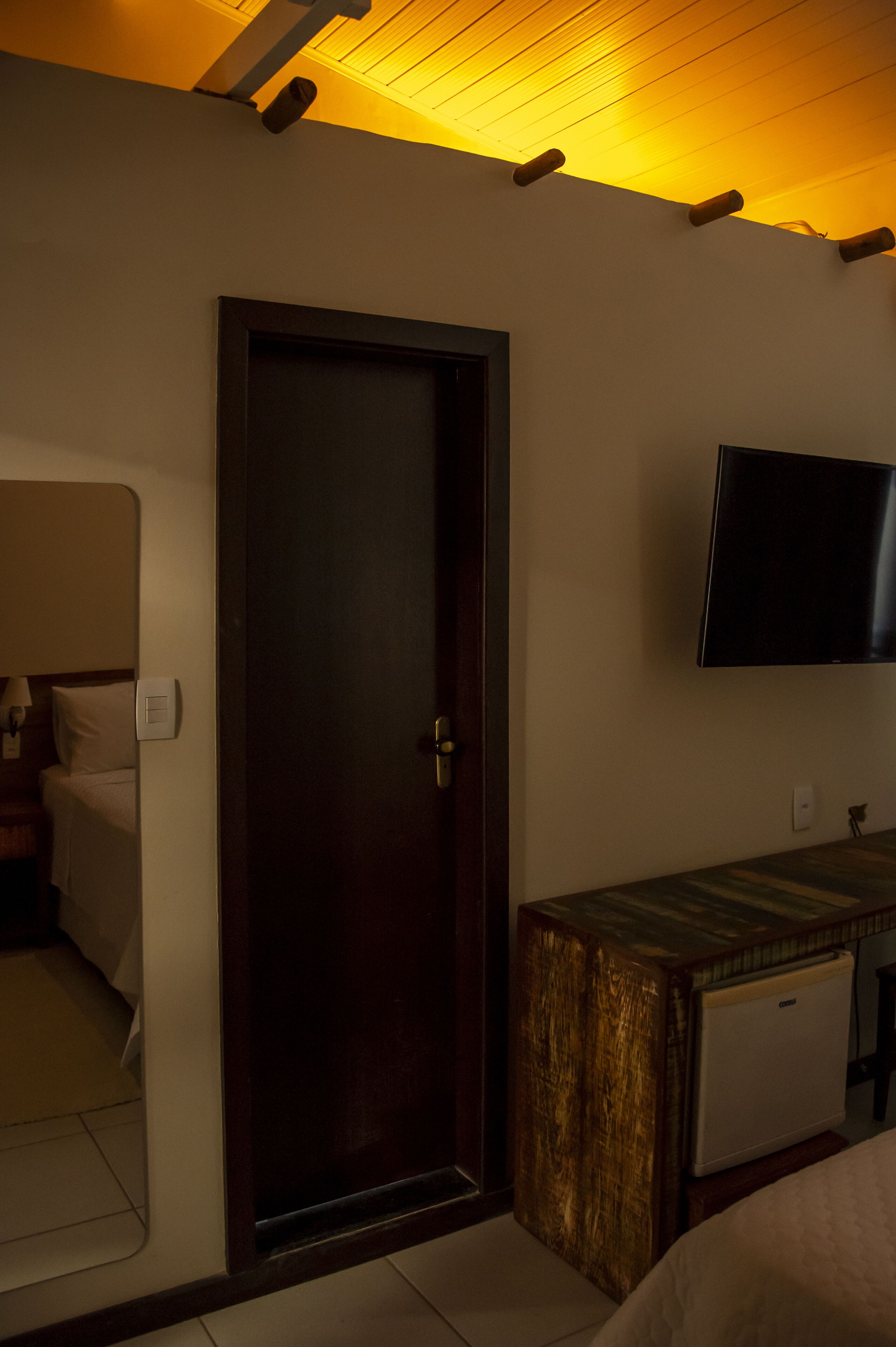 Una habitación de hotel con puerta de madera, cama, mesita de noche, televisor montado en la pared y un mueble pequeño.