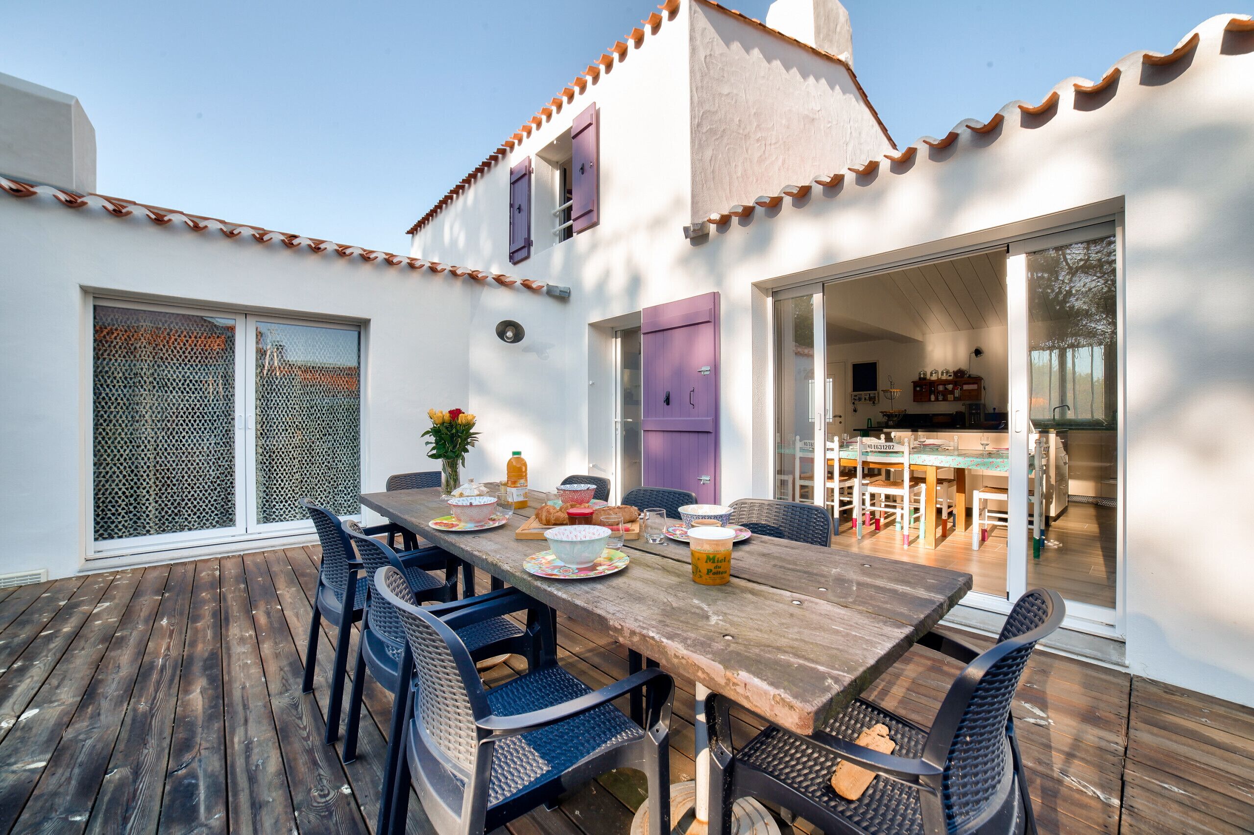 Une terrasse en bois avec une table dressée pour le repas, entourée de chaises, et une vue sur l’intérieur de la maison à travers une porte vitrée.
