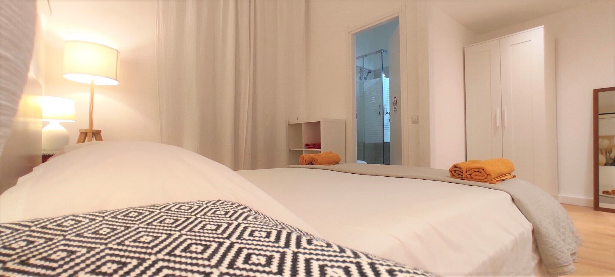 A hotel room with a large bed, a bedside lamp, a wardrobe, and a bathroom visible through a glass door.