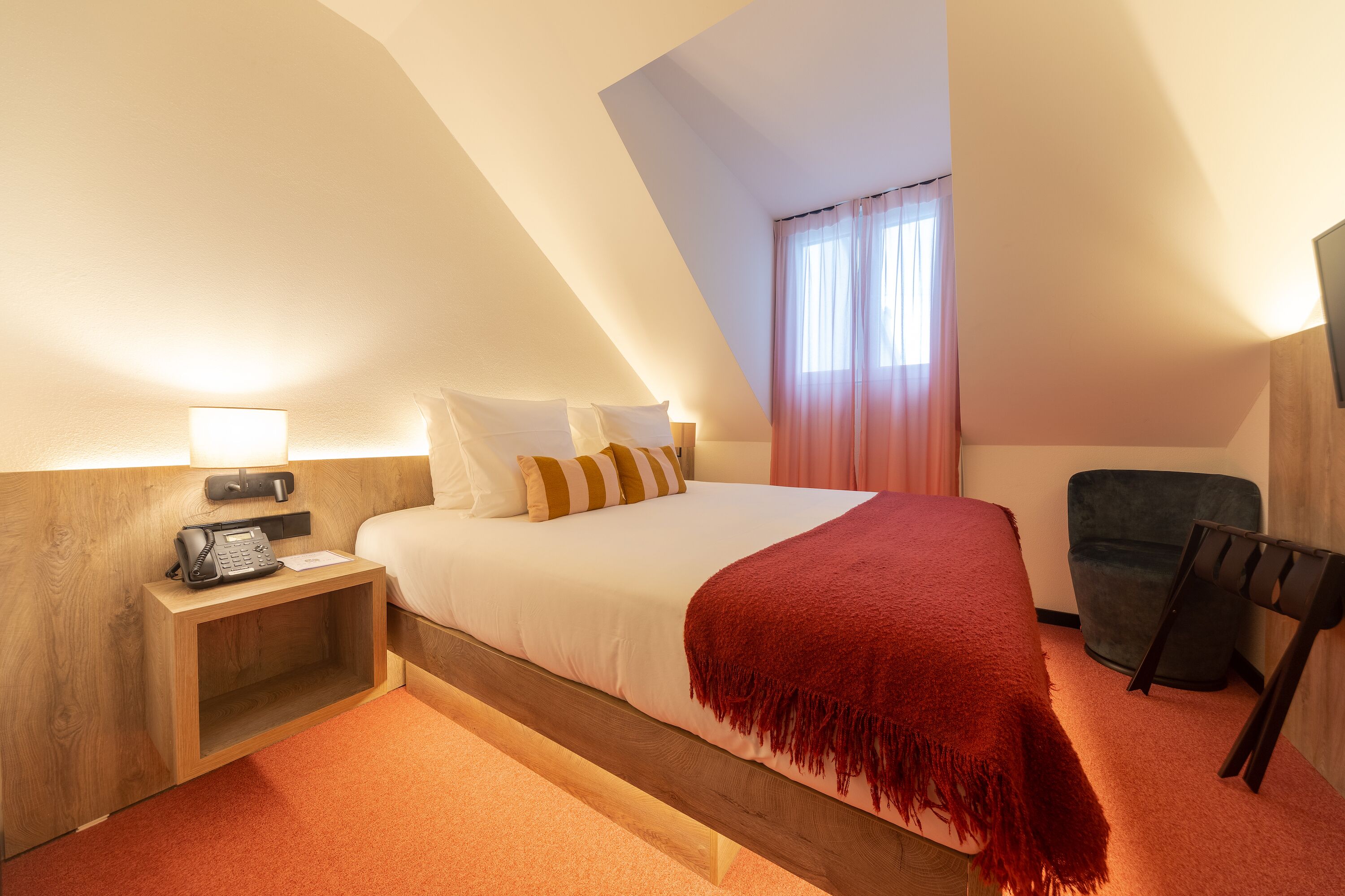 A hotel room with a bed, a nightstand with a phone, a chair, and a window with curtains.