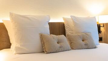 Una cama bien hecha con almohadas blancas y beige, un cabecero de madera y una lámpara fijada a la pared.