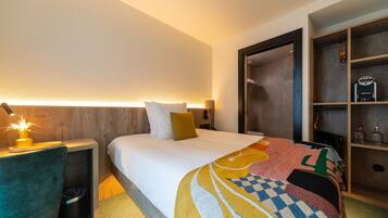 Una habitació d'hotel amb un llit ben fet, una taula de nit amb una làmpada, un gresol i una prestatgeria de fusta amb articles de tocador.