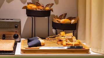 Een keukentafel met een broodrooster, een snijplank met brood en een mes, en twee zwarte standaardjes met brood.