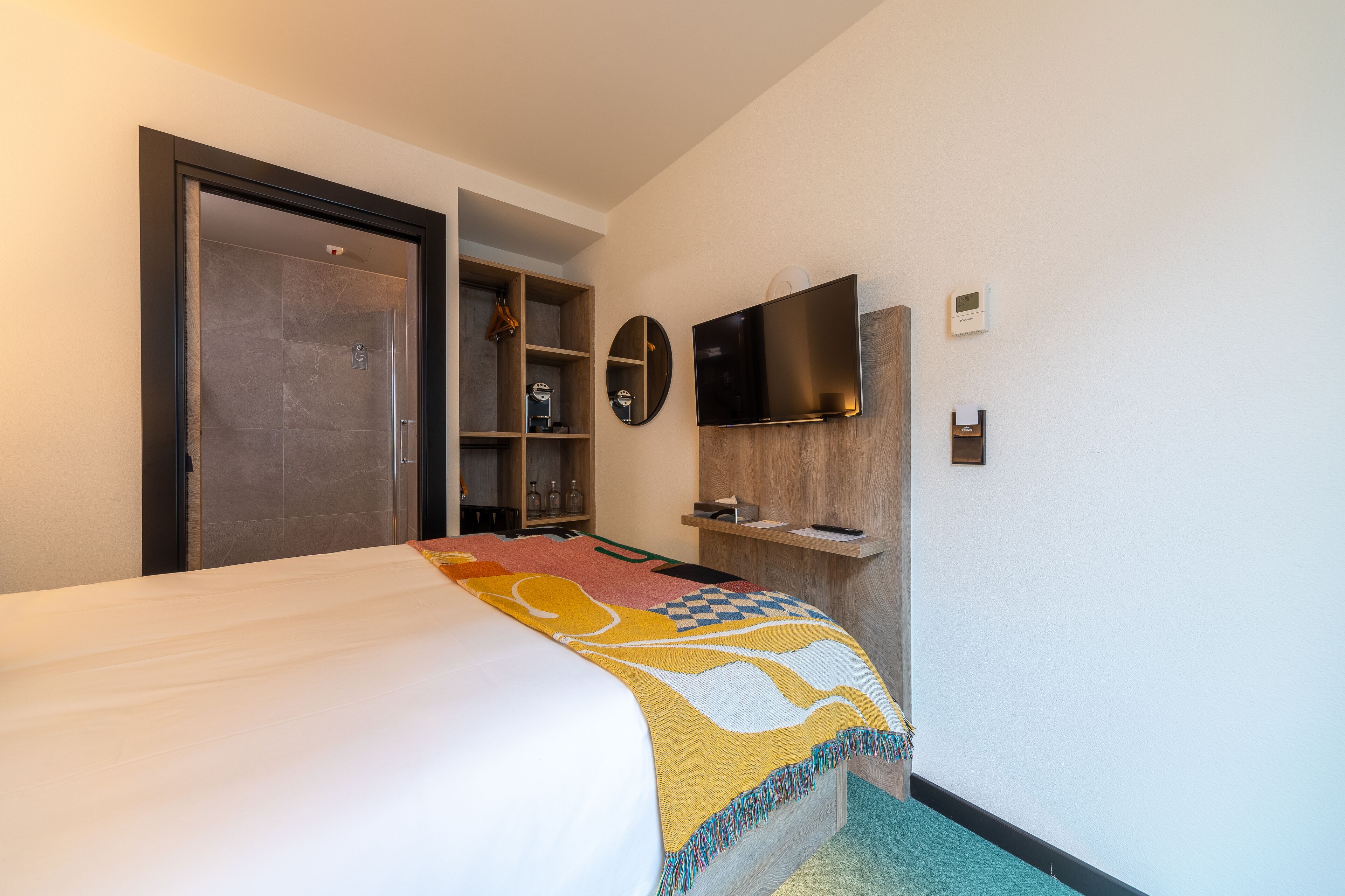A hotel room with a bed, a TV mounted on the wall, a wooden shelf, and a door leading to a bathroom.
