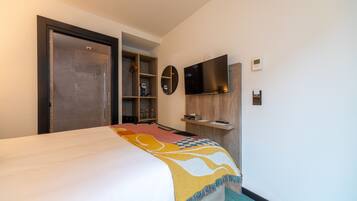 A hotel room with a bed, a TV mounted on the wall, a wooden shelf, and a door leading to a bathroom.