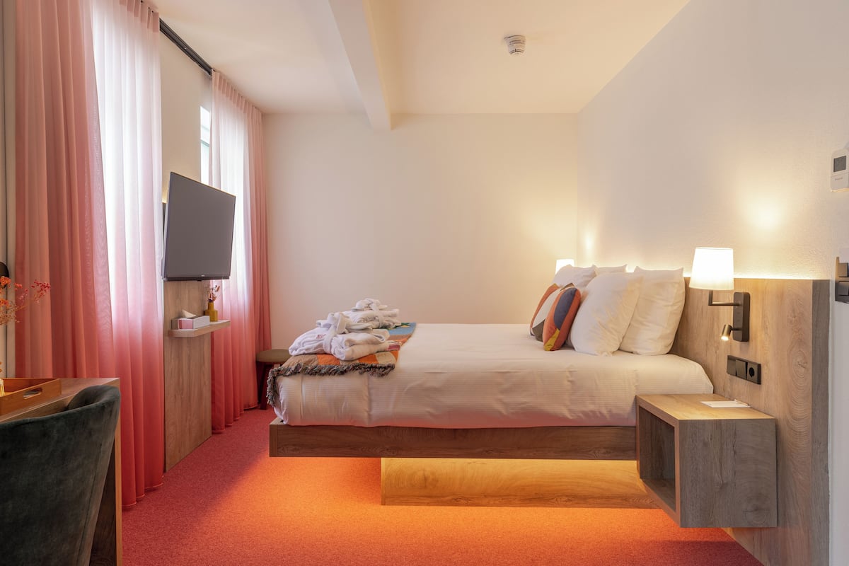 A hotel room with a bed, a TV, a bedside table, a lamp, and a wooden nightstand.