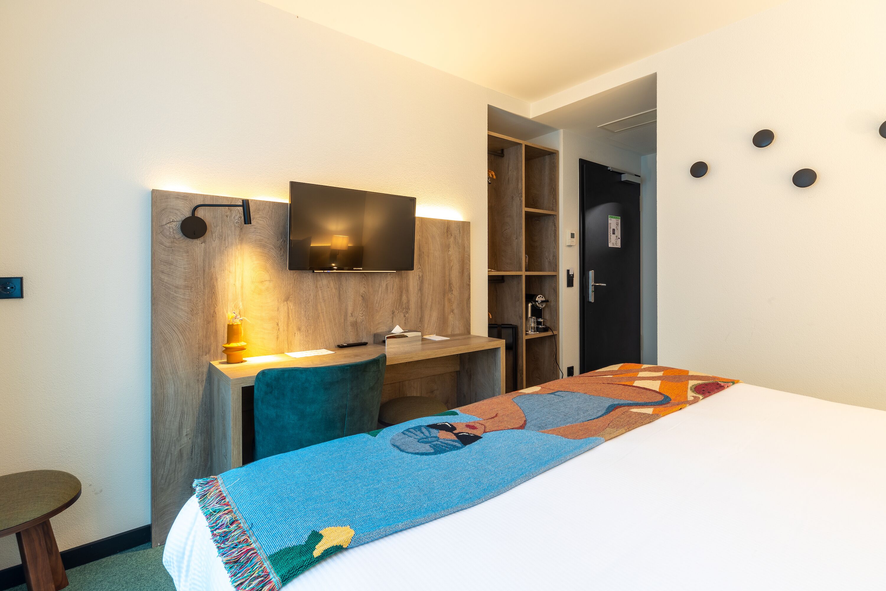 Una habitación de hotel con cama, escritorio, televisor montado en una pared de madera y armario.