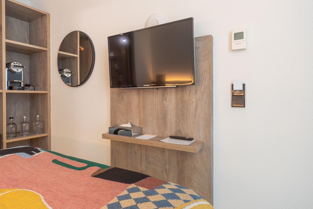 A bedroom with a bed, a TV mounted on the wall, a wooden shelf with toiletries, a mirror, and a thermostat.