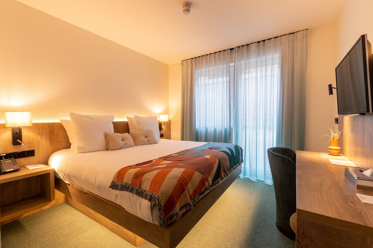 A hotel room with a large bed, bedside lamps, a desk with a flat-screen TV, and a large window with curtains.
