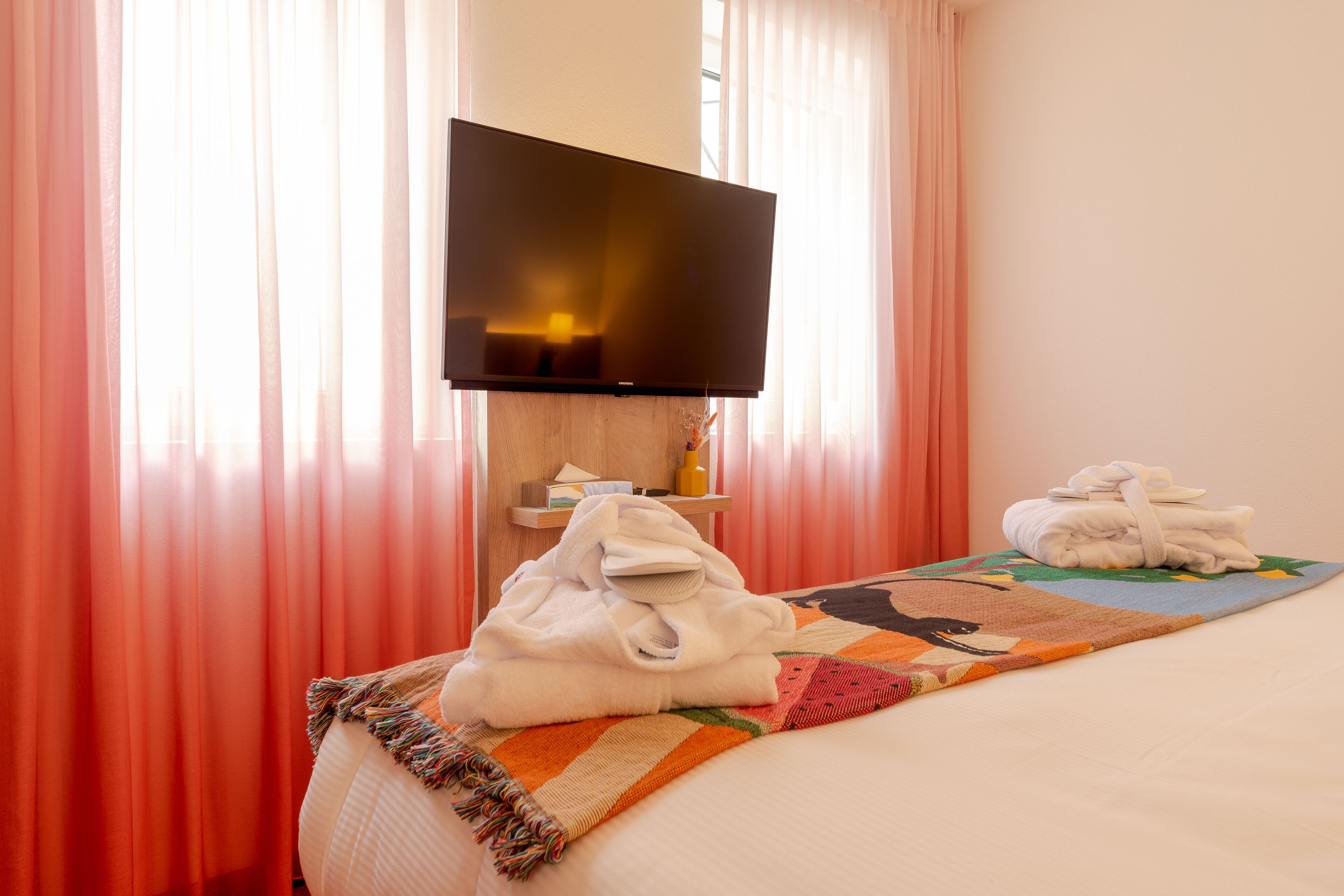 Een hotelkamer met een bed, een flatscreen televisie aan de muur, een kleine tafel met een fles en een doos, en een kleurrijk vloerkleed.