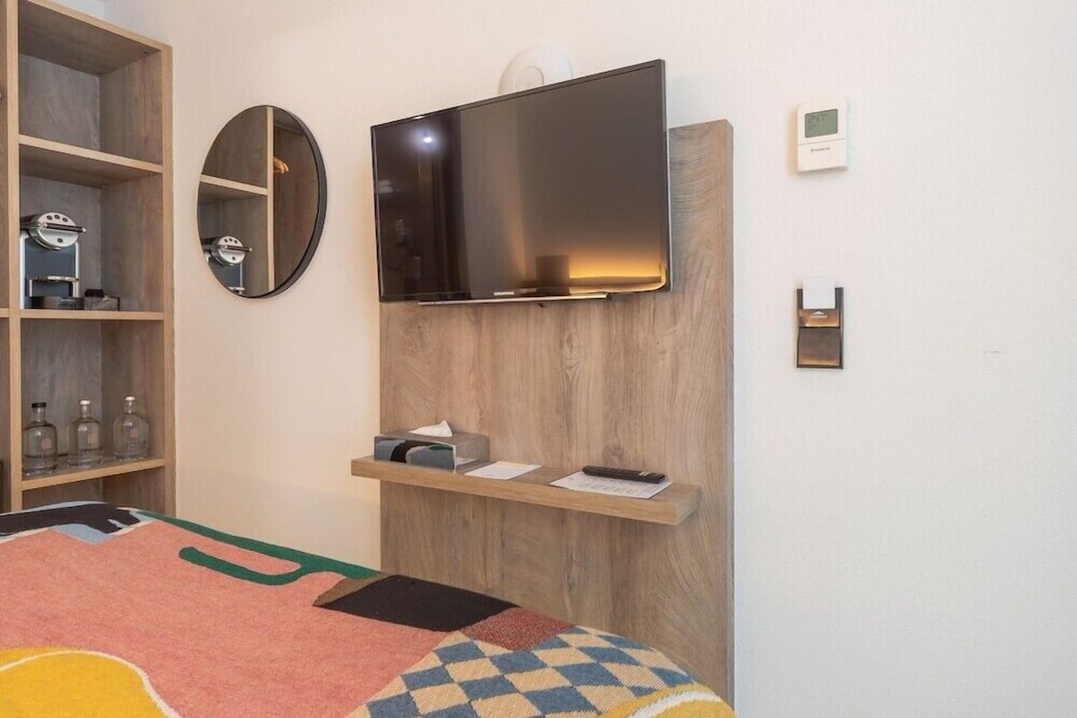 A hotel room with a bed, a mounted TV, a wooden shelf with toiletries, a mirror, and a thermostat.