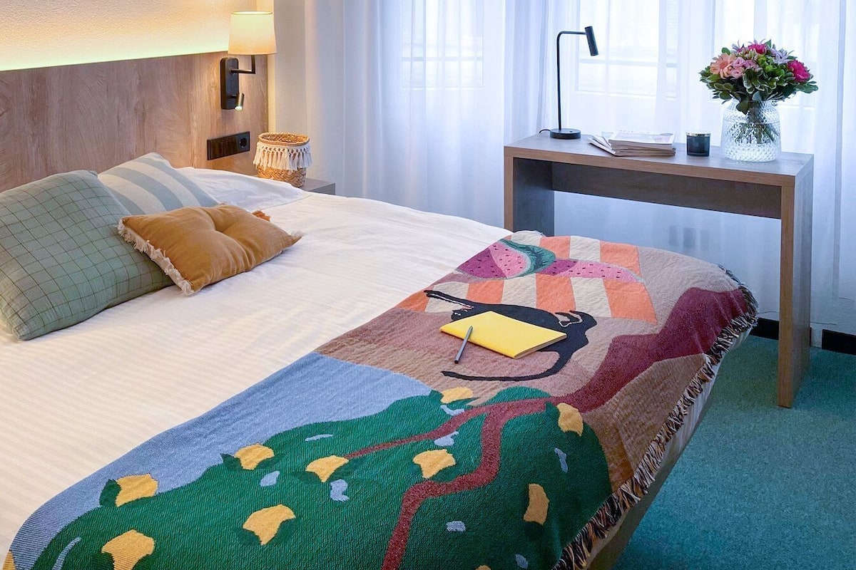 A hotel room with a bed, a desk, a lamp, and a vase with flowers.