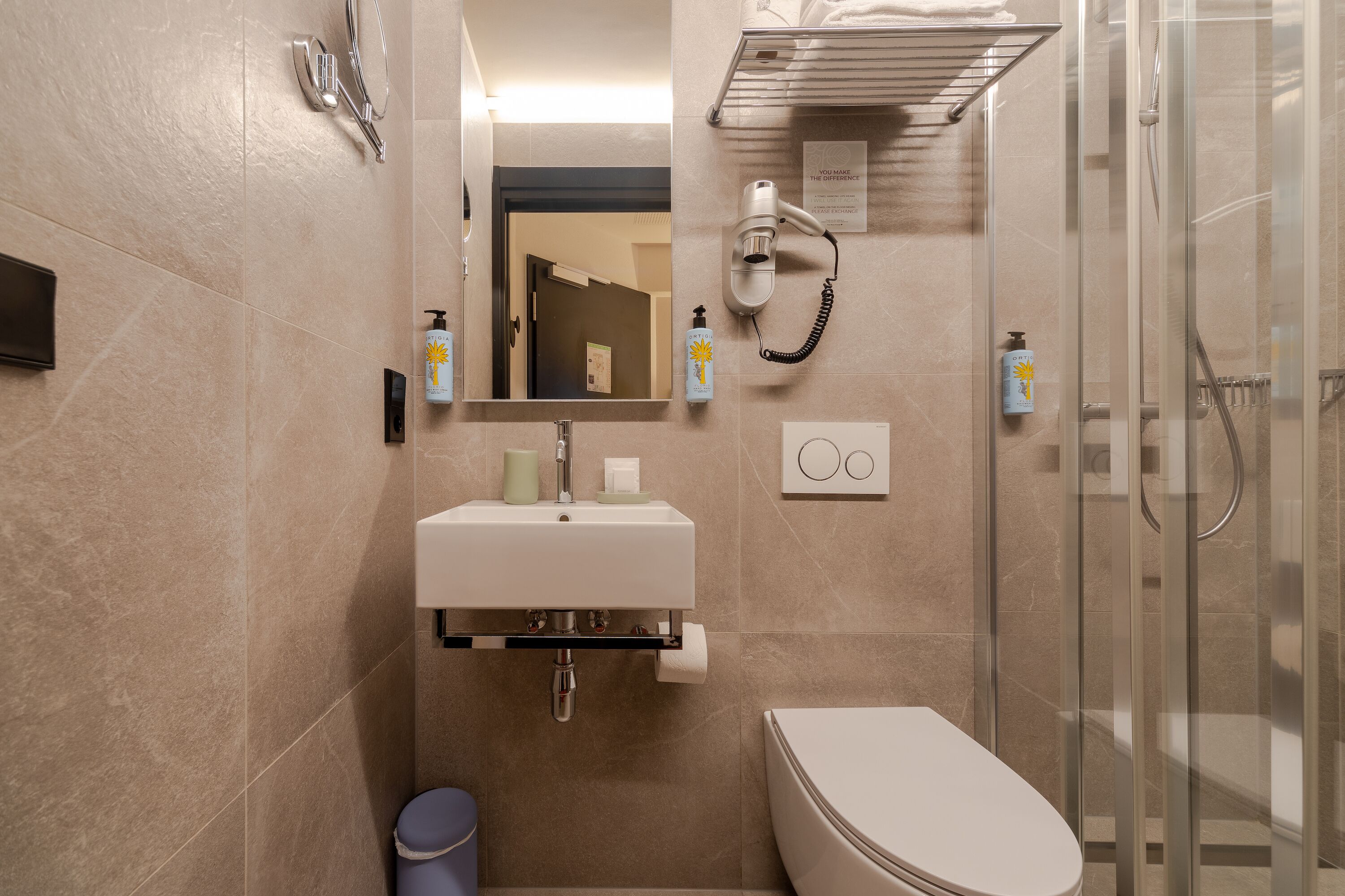 Ein modernes Badezimmer mit Waschbecken, Spiegel und Toilette. Ein Föhn ist an der Wand angebracht und ein Duschbereich ist sichtbar.